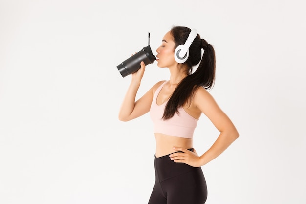 Sport, wellbeing and active lifestyle concept. Profile portrait of sexy asian fitness coach, woman in headphones drinking water from bottle during workout in gym, standing white wall