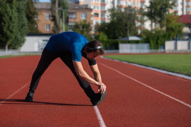 Sportive young man wears headphones doing stretching at the running track. Empty space