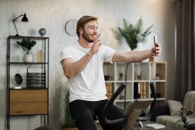 Sports bearded caucasian man in sportswear having online video call while cycling bike