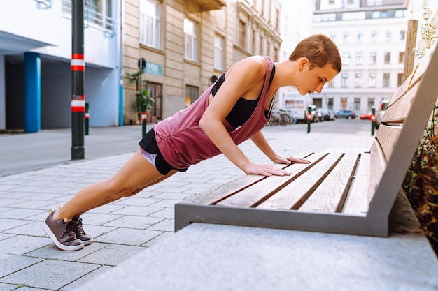 sports girl teenager does push-ups on arms bent at elbows from bench on city street in sun