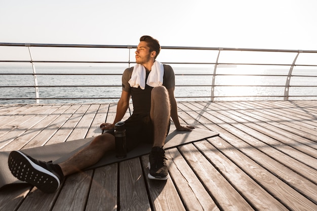 Sports man outdoors on the beach sitting near bottle with water on rug