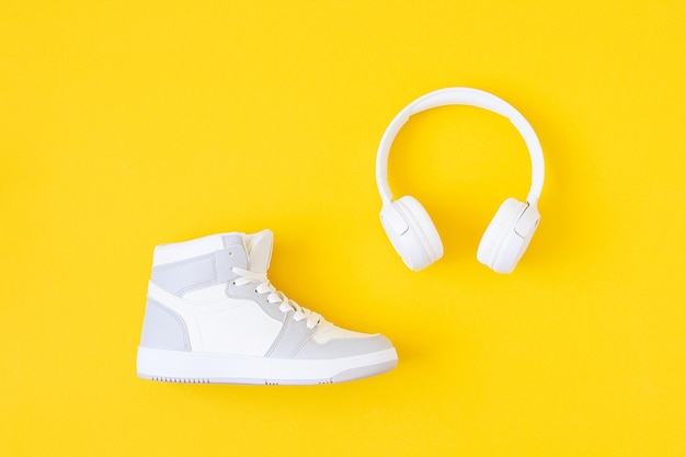 Sports shoe sneakers and white headphones on yellow background Music audio books learning during sports leisure activity concept Top view Flat lay