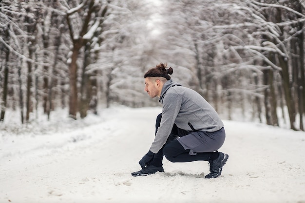 Sportsman crouching in snow and tying shoelace at winter. Sportswear, winter fitness, healthy life