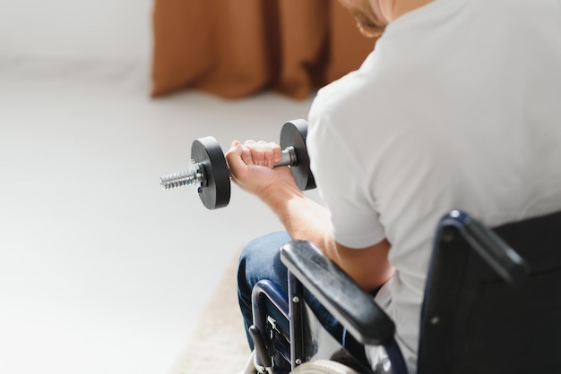 Sportsman sitting in wheelchair and outstretching arms with dumbbells during rehabilitation exercise in modern medical center Man sitting on wheelchair at home