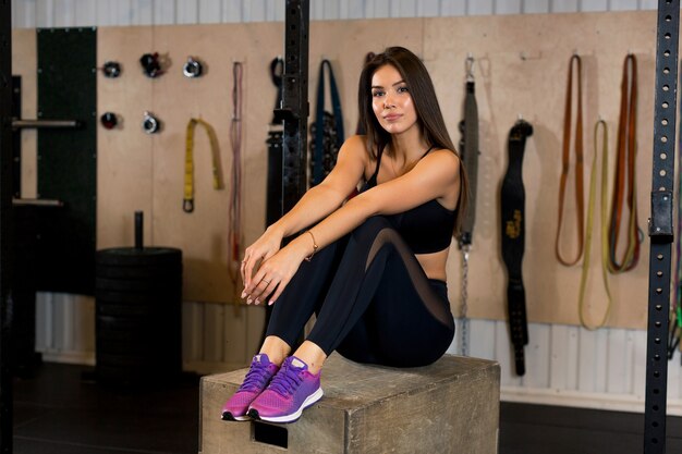 A sportswoman in black uniform and pink sneakers sits on a wooden box in the gym against a backdrop of sports equipment