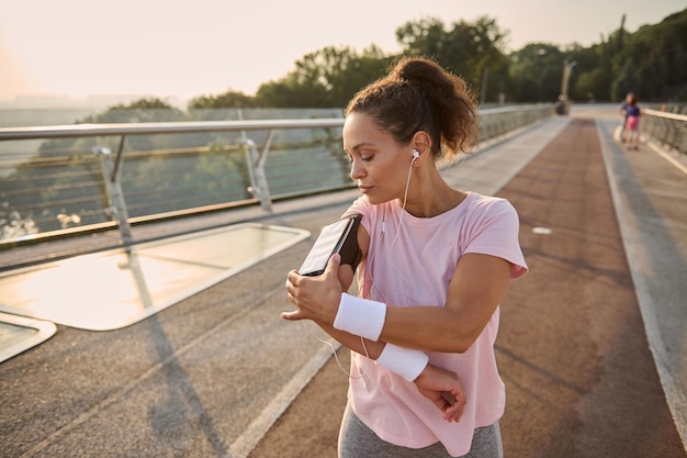 Sportswoman, female athlete runner adjusting mobile applications on her smartphone in a holder on her hand while exercising outdoor, standing on a treadmill on the city bridge.