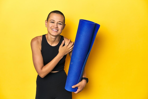 Sportswoman holding yoga mat yellow studio background laughing and having fun