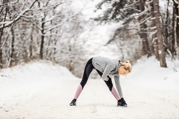 Sportswoman standing on snowy path in nature at winter and doing stretching and warmup exercises. Healthy life, winter fitness, outdoor fitness