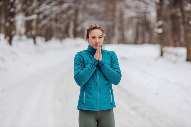 Sportswoman trying to warm up hands on snowy weather while standing in nature. Chilly weather, outdoor fitness, nature, winter fitness