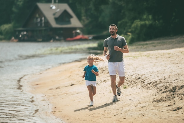 Sporty as father. Full length of cheerful father and daughter in sports clothes jogging at the riverbank together