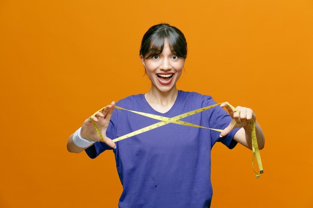 Sporty beautiful woman in sportswear holding measure tape looking at camera happy and positive smiling cheerfully standing over orange background