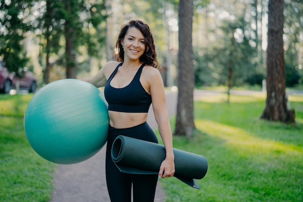 Sporty European woman in cropped top and leggings carries rolled up karemat and fitness ball going to have aerobics exercises poses outdoor against green nature background has pilattes class