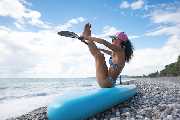 A sporty girl in a swimsuit with a paddle poses sexually with a sup board in summer against the background of the sea