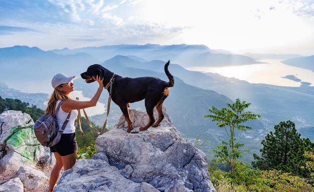 A sporty girl with a backpack stands on the edge of a mountain with a rottweiler dog