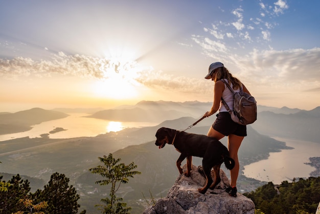 A sporty girl with a backpack stands on the edge of a mountain with a rottweiler dog