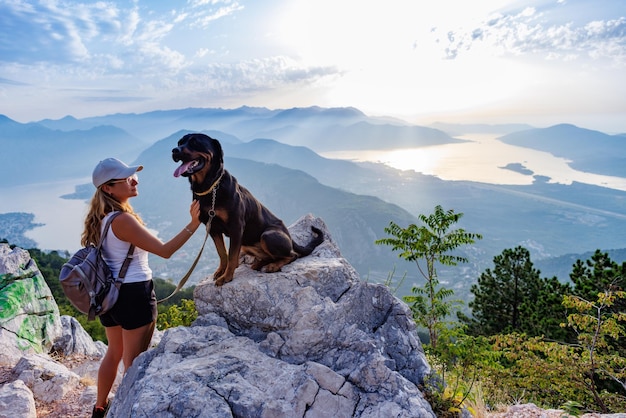 A sporty girl with a backpack stands on the edge of a mountain with a rottweiler dog