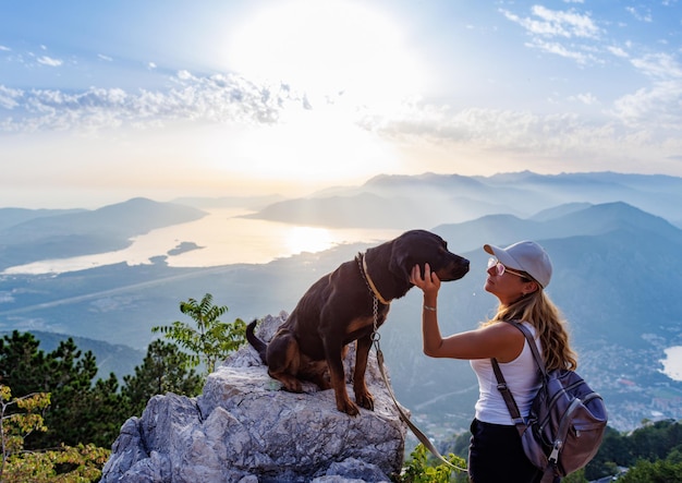 A sporty girl with a backpack stands on the edge of a mountain with a rottweiler dog