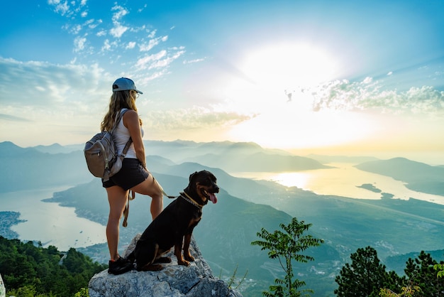 A sporty girl with a backpack stands on the edge of a mountain with a rottweiler dog