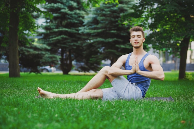 Sporty guy practicing yoga sitting on grass in park. Young man meditating with closed eyes, namaste on green grass in park, copy space