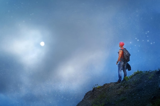 Sporty guy with a backpack on the edge of the cliff against a blue sky with stars Travel and freedom concept