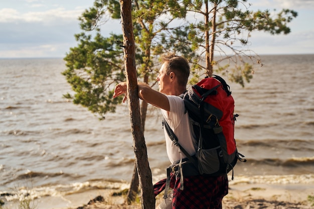 Sporty male with backpack is standing in beach and leaning against tree while enjoying view