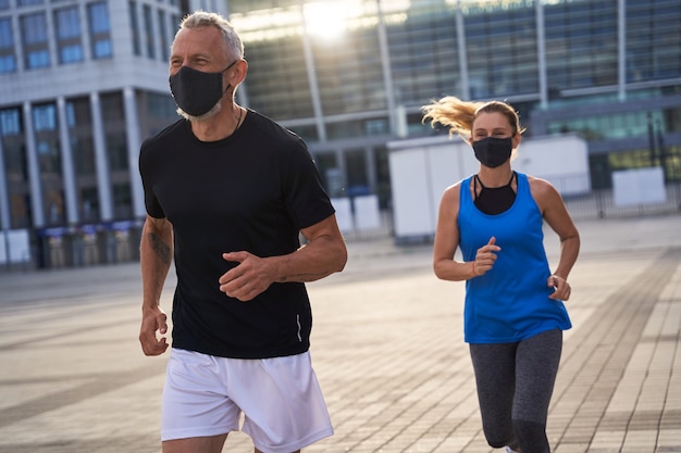 Sporty middle aged couple wearing protective masks running in urban environment staying active