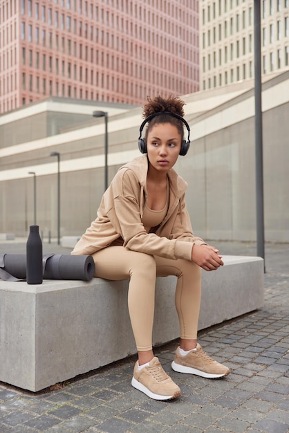 Sporty tired woman dressed in sportswear listens music from playlist via headphones poses outdoors near rolled fitness mat and bottle concentrated aside exercises in urban setting Healthy lifestyle