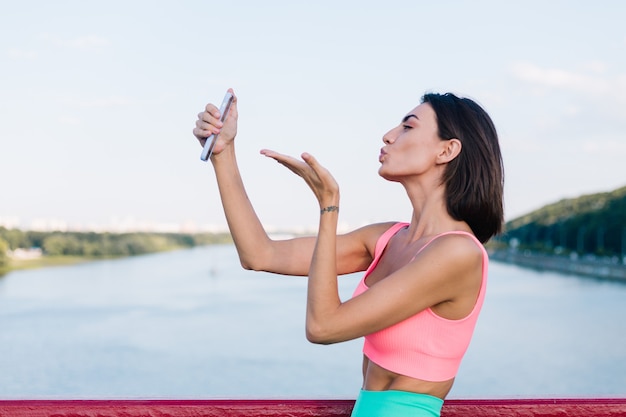 Sporty woman in fitting sport wear at sunset at modern bridge with river view happy positive smile with mobile phone take photo selfie video for socials stories