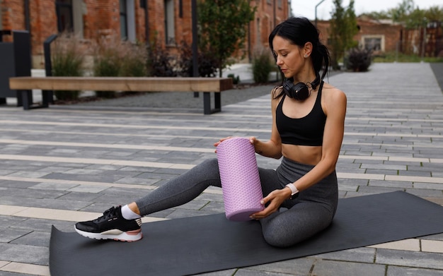 Sporty woman with a massage roller in hands sits on a mat on the street