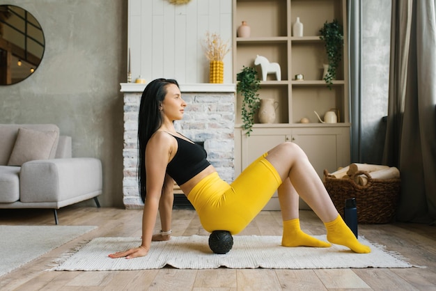 Sporty woman in yellow and black sportswear a bra and bicycles practicing at home or in a yoga studio rolling an mfr ball or a massage ball on her buttock