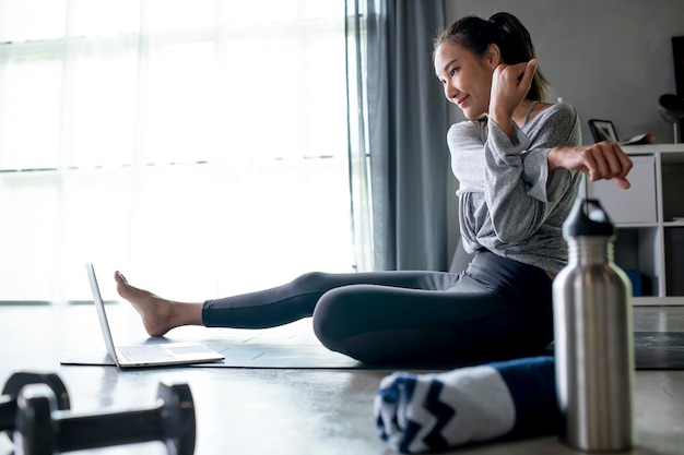 Sporty young Asian woman exercising at home watching fitness video on Internet or having online fitness class using laptop living room interior copy space