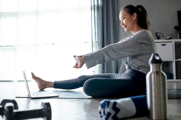 Sporty young Asian woman exercising at home watching fitness video on Internet or having online fitness class using laptop living room interior copy space