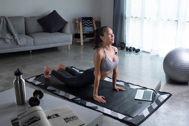 Sporty young Asian woman planking in front of laptop living room interior side view copy space Healthy young lady in sportswear exercising at home watching online fitness class