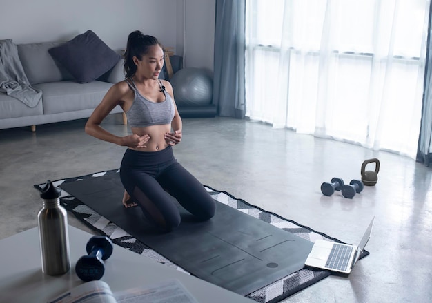 Sporty young Asian woman in sportswear exercising at home watching fitness video on Internet or having online fitness class using laptop living room interior copy space