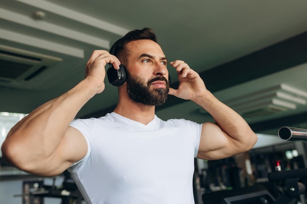 Sporty young man putting headphones into his ears while resting after his workout in gym