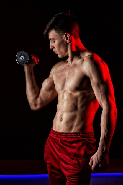 Sporty young man training with dumbbells against dark background