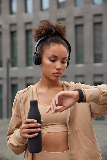 Sporty young woman checks results of fitness training holds bottle with fresh water dressed in activewear listens music via headphones poses outdoors against blurred buiding. Healthy lifestyle