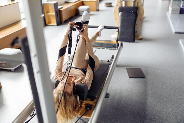 Photo sporty young woman putting on bands of a pilates reformer machine bed while getting ready for workout in gym studio healthy lifestyle concept