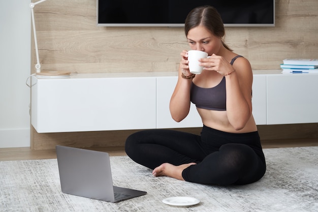 Sporty young woman sit on floor in living room practice yoga on online computer lesson female