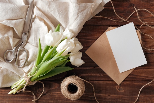 Spring bouquet of white tulip flowers, blank paper card, scissors, twine on rustic wooden desk.