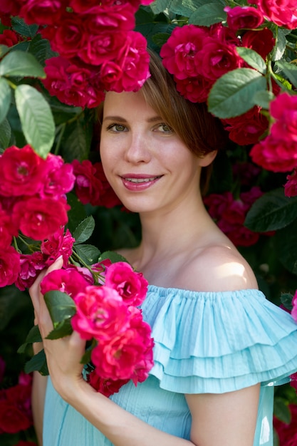 Spring. Close up portrait of a happy pretty redhead girl with charming smile wearing trendy blue light dress on a nature of blooming roses. Outdoor.