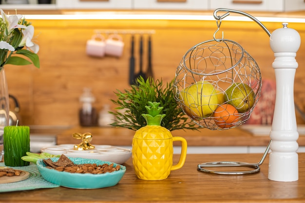 Spring counter with fresh fruit, almond in platter and pineapple teapot, flowers in vase, candle, fruit in round-shaped hanger and mill of pepper.