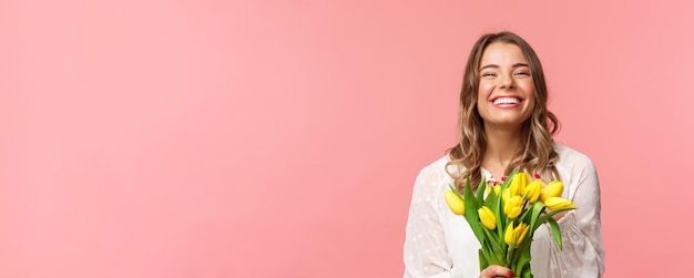 Spring happiness and celebration concept Closeup of happy and carefree blond european girl receive beautiful bouquet of flowers holding yellow tulips smiling and laughing pink background