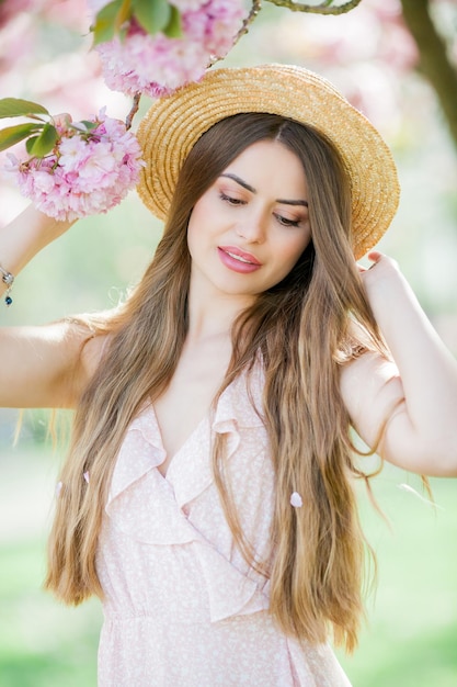 Spring portrait of a young beautiful woman in pink blossoms Young beautiful model with long hair in a wicker hat and a light dress near the cherry blossoms