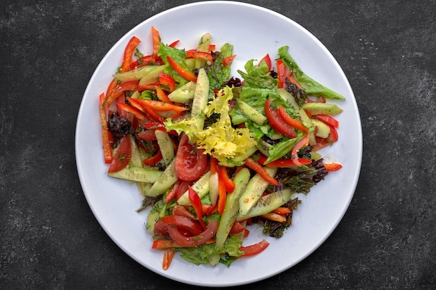 Spring salad of fresh vegetables, cucumbers, tomatoes, lettuce, bell pepper on a white plate, on a dark background