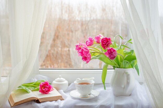 A spring still life a bouquet of tulips in a vase a cup of tea and an old book are lying on window