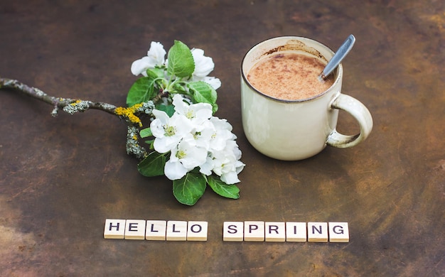 Spring still life on a dark background. The inscription "Hello spring" A cup of hot cocoa, a blooming branch of an apple tree. Vintage style photo, flower arrangement with a branch