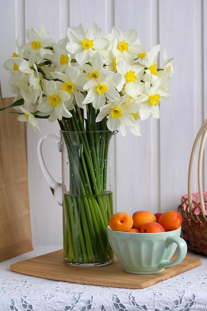 Spring still life with a bouquet of white daffodils in a glass jug and a bowl of apricots