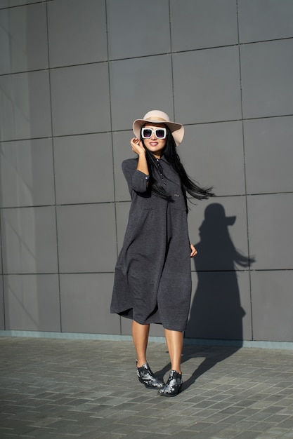 Spring trendy collection. Happy beautiful brunette girl wearing stylish grey dress and fashionable hat. Model in trendy outfit posing on grey wall