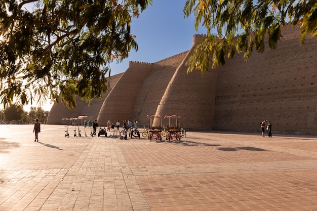 Square around ancient Ark Citadel with walking people and rental bikes at sunset Bukhara Uzbekistan